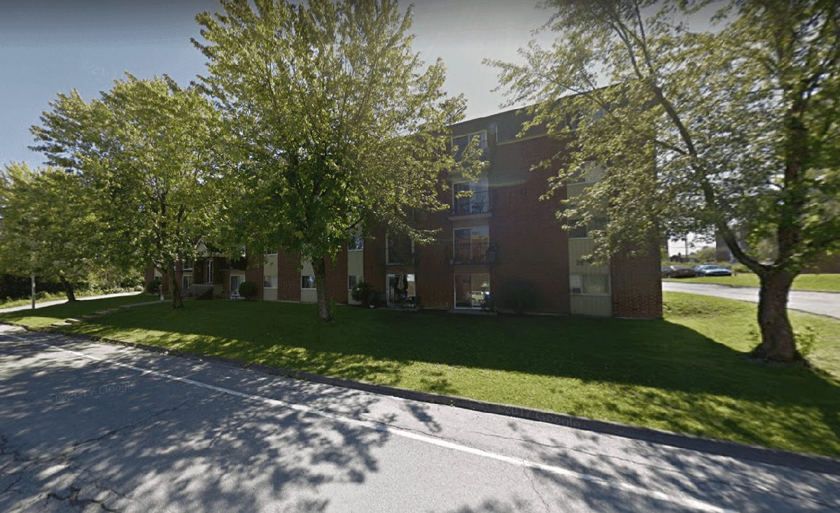 Brown brick apartment building surrounded by mature trees, seen from the street