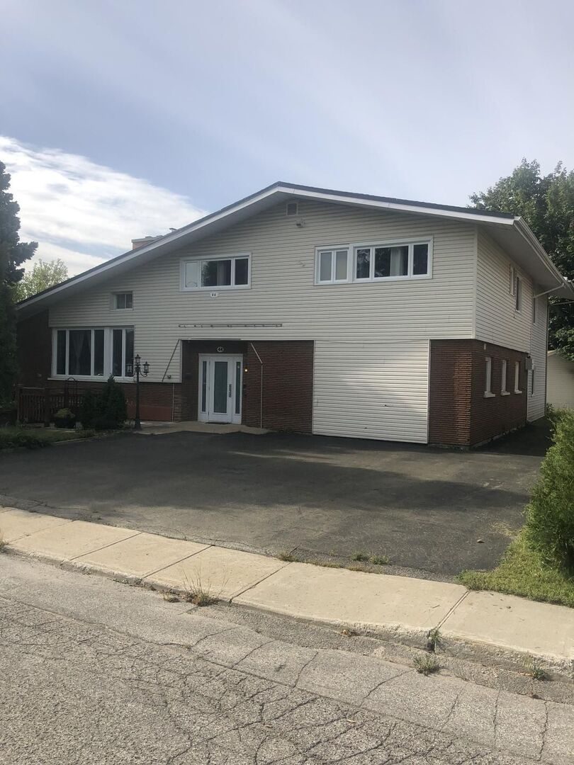 Two-storey building with white siding, a brick base and a large paved parking area